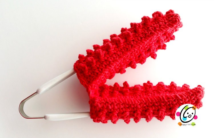 A red, textured crochet hanger cover with a zigzag pattern adds charm to your spring cleaning routine. With the metal hanger peeking through and white handles, it's both stylish and practical. The image features the Gooyer logo in the bottom right corner. -Marly Bird