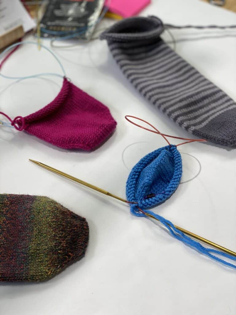 Four knitted sock pieces, including pink, blue, multicolored, and gray with stripes, are laid out on a table. Each piece is expertly crafted using a toe-up knit sock pattern with circular knitting needles attached. -Marly Bird