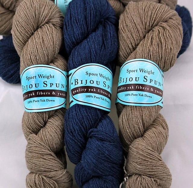 Three skeins of Bijou Spun sport yarn—two light brown, one dark blue—100% yak down with blue labels, ideal for knit shawl projects.