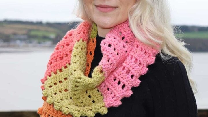 A woman models a crochet scarf in pink, yellow, and orange with visible stitch detail over a black sweater outdoors.