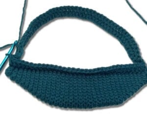 A crochet oval in dark teal yarn, partially completed, with the hook still attached on a white background.