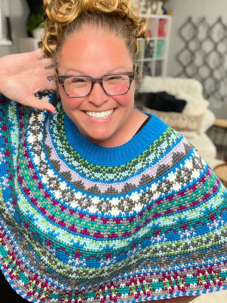 A smiling woman with curly hair and glasses wears a colorful crochet swoncho. She poses with one hand behind her head in a bright, cozy room with shelves and a white chair in the background. Perfect for the Prism Make-Along 2025!. -Marly Bird