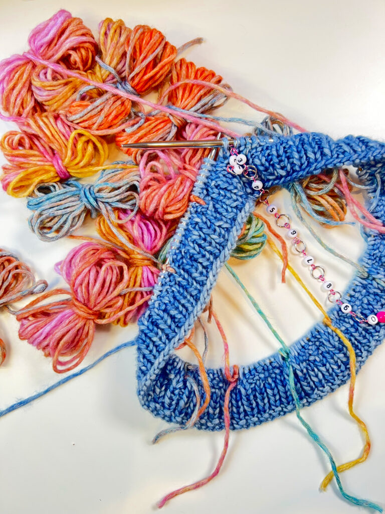 knitting work in progress, image shows Caron Blossom Cake in denim color on knitting needles with bobbins of other colors dangling from the project - also includes a row counter - Marly Bird