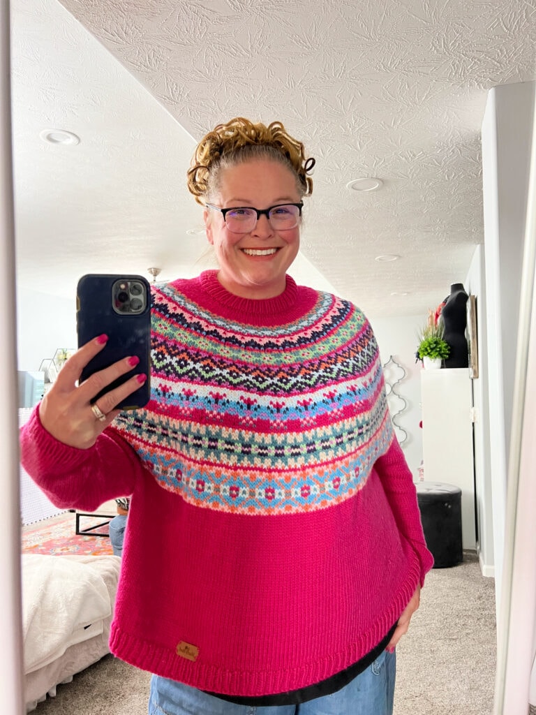 A smiling person with curly hair, glasses, and a bright pink Prism Make-Along sweater with a colorful patterned yoke takes a selfie in a well-lit, cozy room with carpet and modern decor. -Marly Bird