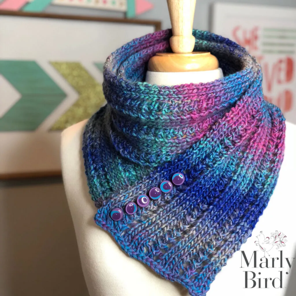 A mannequin displays a textured, multicolored Rib and Lace Knit Scarf Cowl in shades of blue, purple, and pink, fastened with blue buttons. The Marly Bird logo appears in the lower right corner. Geometric wall art is in the background. -Marly Bird
