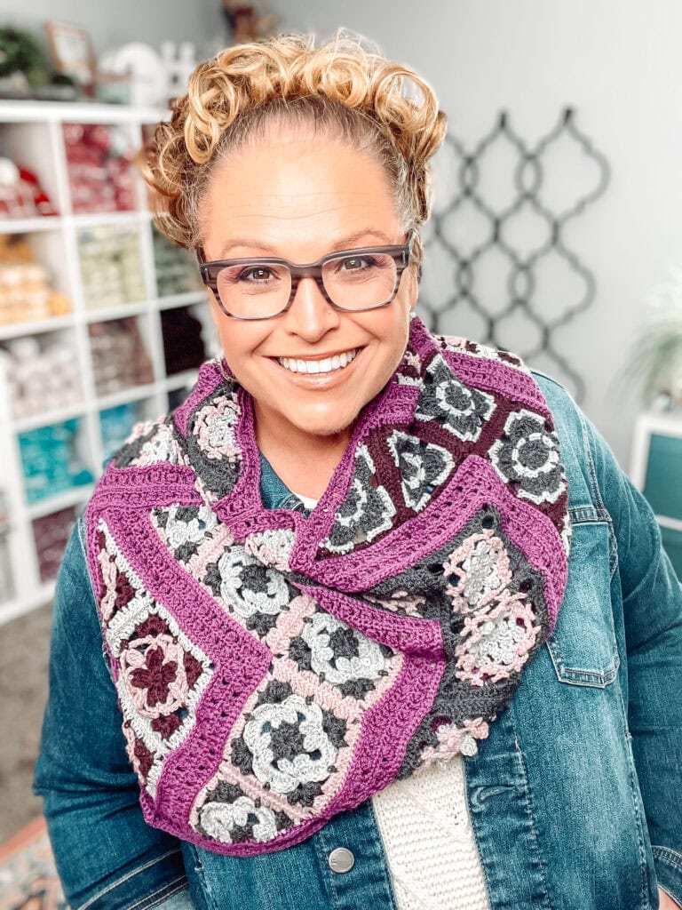 A smiling person with curly hair and glasses wears a purple, gray, black, and pink crocheted scarf over a denim jacket, standing in a cozy room with yarn-filled shelves and geometric wall decor. -Marly Bird