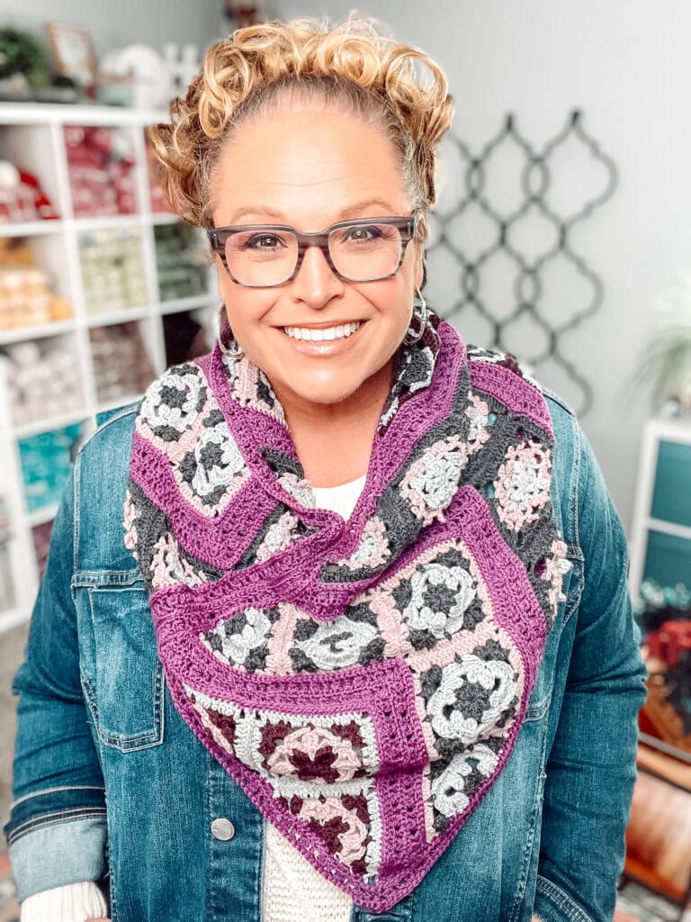 Smiling woman with curly hair and glasses wearing a denim jacket and a colorful, crocheted scarf with purple, pink, and gray patterns, standing in a room with yarn shelves in the background. -Marly Bird