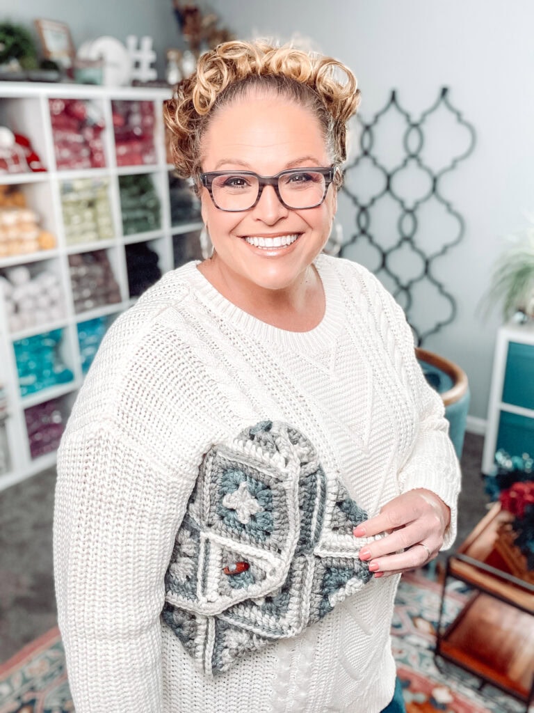 A smiling woman in a white sweater and glasses holds a gray and white geometric-patterned crocheted bag made with a One Ball Crochet Granny Square Pattern. Behind her are shelves filled with yarn and a colorful rug on the floor. -Marly Bird