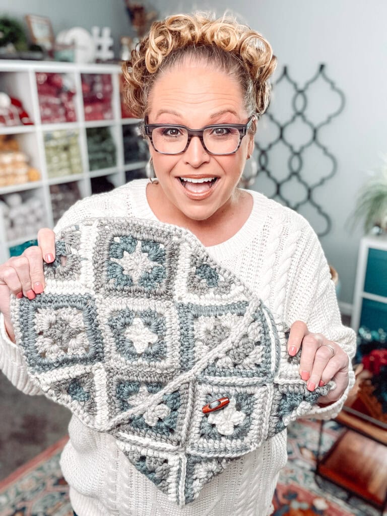 A smiling woman with glasses and curly hair holds up a crocheted item made of gray, white, and green granny squares. She is wearing a white sweater in a cozy, yarn-filled room. -Marly Bird