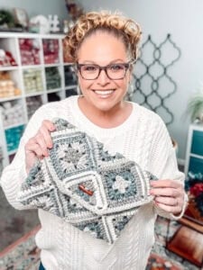 Woman displays a granny square accessory in gray, white, and green Tunisian crochet inside a cozy craft room.