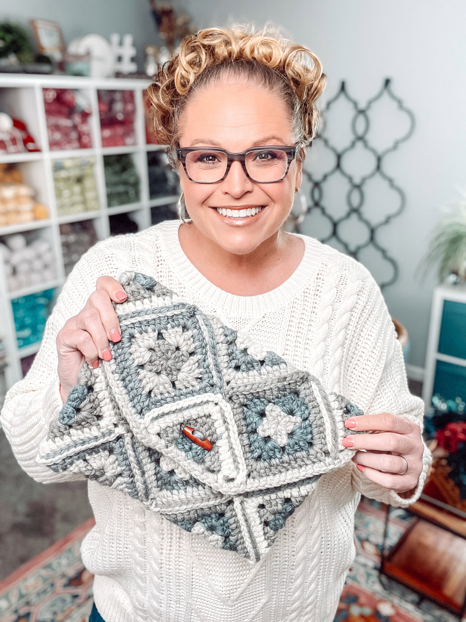 A woman wearing glasses and a white sweater smiles while holding a crocheted blanket made with the One Ball Crochet Granny Square Pattern. Shelves with colorful yarn and home decor are visible in the background. -Marly Bird