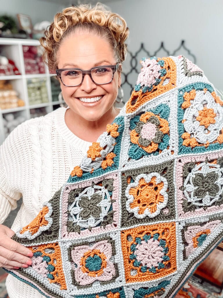 A smiling woman with glasses and curly hair holds up a colorful, handmade crochet blanket featuring intricate floral and geometric patterns in orange, green, gray, and cream. Shelves with yarn are visible in the background. -Marly Bird
