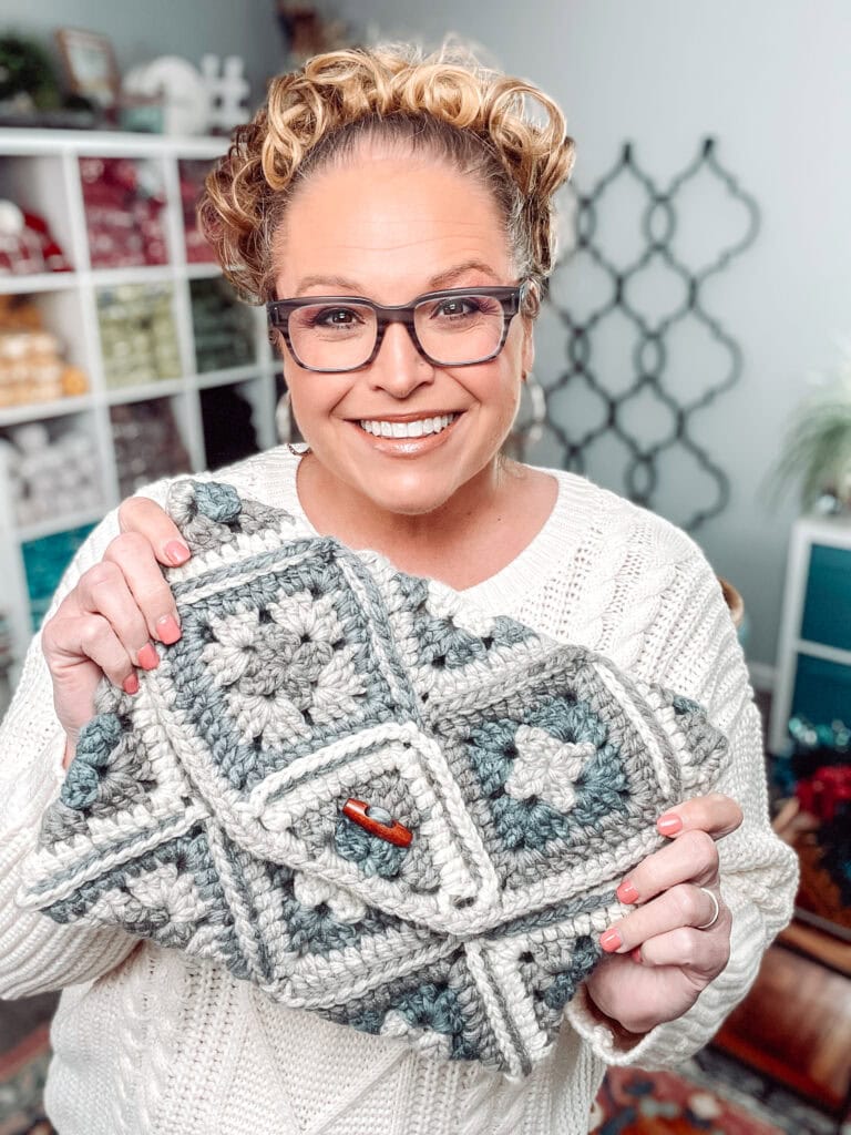 Smiling woman with glasses and curly hair holds a handmade crochet bag made of gray and white granny squares. She wears a white sweater, and yarn shelves are visible in the background. -Marly Bird