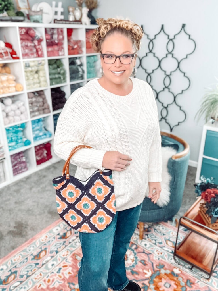 A smiling woman wearing glasses, a white sweater, and jeans stands indoors, holding a colorful crochet bag. Behind her are shelves filled with yarn and a cozy, decorated room with a patterned rug and chair. -Marly Bird