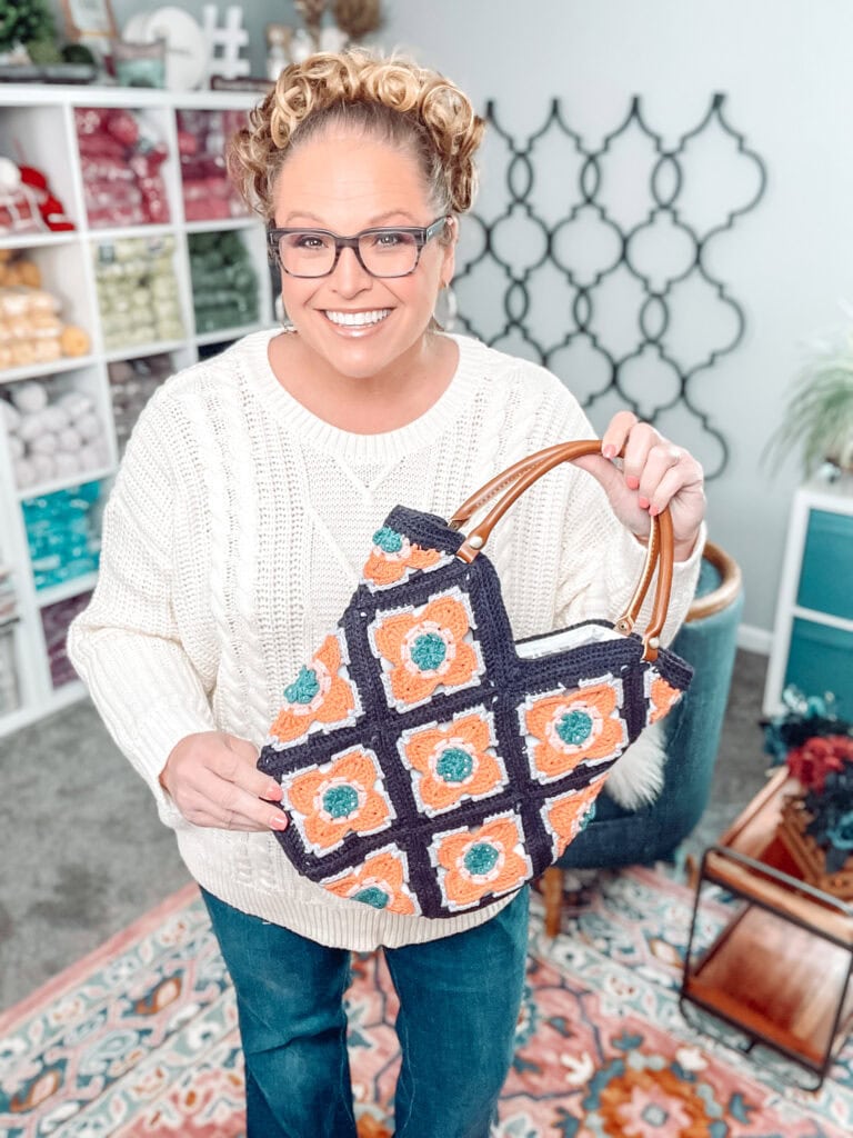 A smiling woman with glasses and curly hair stands indoors, holding a crocheted handbag with a colorful square pattern. She wears a white sweater and jeans, and yarn shelves are visible in the background. -Marly Bird