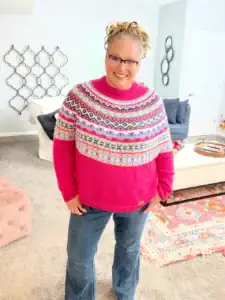 A smiling woman with glasses stands in a cozy living room, wearing a bright pink, patterned sweater made from free knitting and crochet patterns. The room features light walls, a cream sofa, and colorful decor, including a patterned rug. -Marly Bird