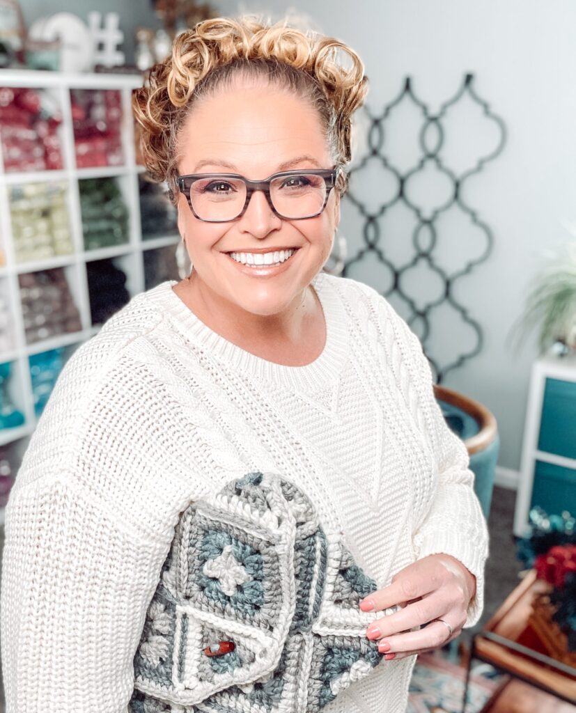 A smiling woman with glasses and curly hair wears a white sweater and holds a crocheted blanket. She stands in a cozy room with cubby shelves of yarn and craft supplies, surrounded by free knitting and crochet patterns. -Marly Bird