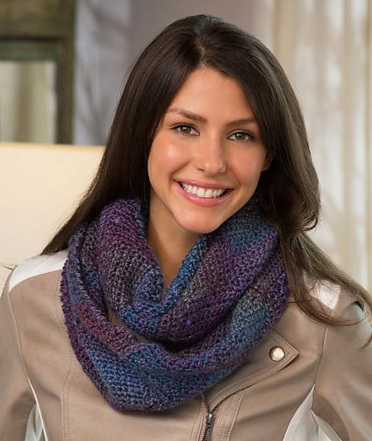 A woman models a crochet infinity scarf in purple and blue hues, showing its textured wrap pattern against a beige jacket.