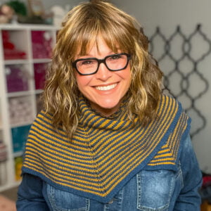Woman models a knit shawl with yellow and blue stripes, showing its cozy textured stitches; denim shirt worn underneath.