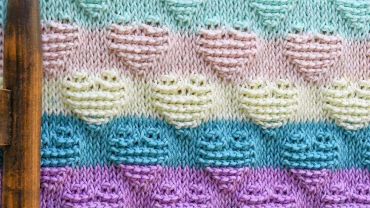 Pastel heart motif crochet blanket with rows of hearts in purple, blue, green, and cream on a wooden rail outdoors.