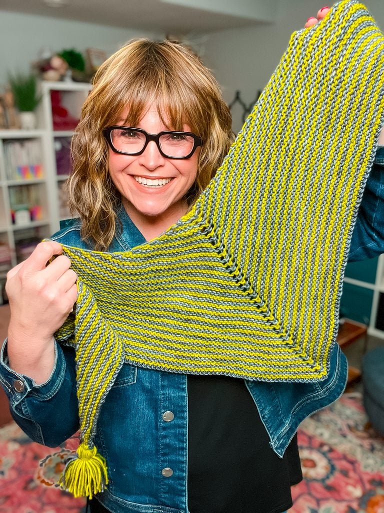 A woman holds up the Super Simple Two Ball Shawl to show its triangular shape, with yellow-green and gray alternating stripes and a bright yellow tassel at the tip. -Marly Bird