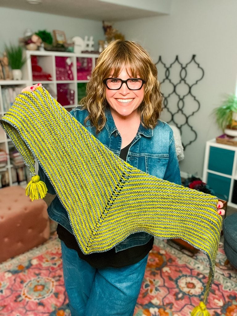 A woman stretches the Super Simple Two Ball Shawl wide to show its full wingspan, displaying the alternating yellow-green and gray garter stitch stripes and triangle construction. -Marly Bird