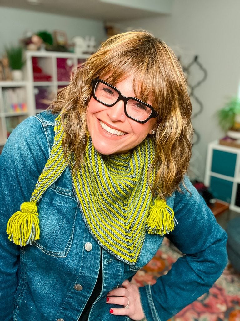 A smiling woman wearing the Super Simple Two Ball Shawl as a cozy neck wrap in bright yellow-green and gray garter stitch stripes with yellow yarn tassels, paired with glasses and a denim jacket. -Marly Bird