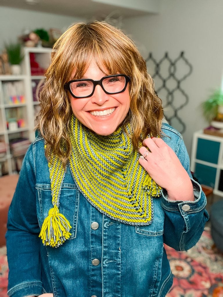 A smiling woman wearing the Super Simple Two Ball Shawl styled as a triangle scarf around her neck, showing the yellow-green and gray striped garter stitch fabric with tassels draped in front. -Marly Bird