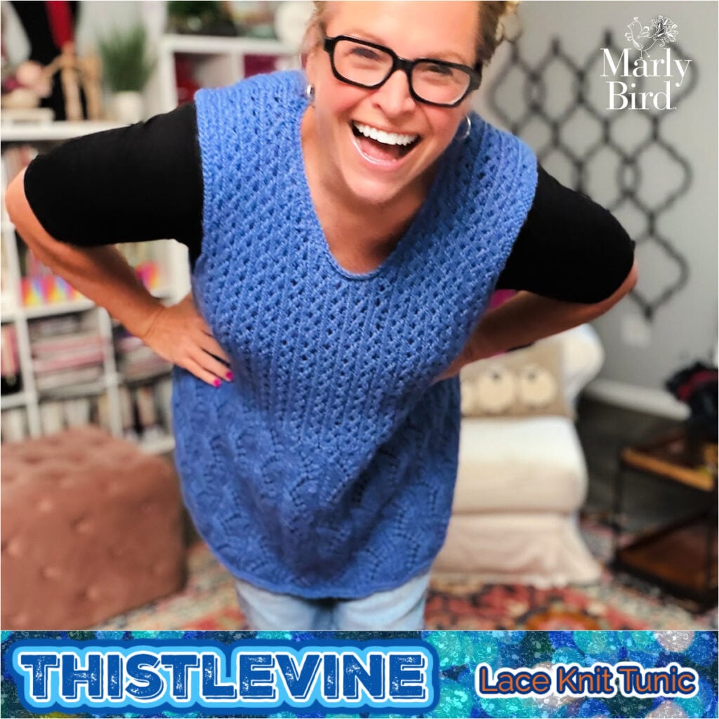 A person models a blue lace knit tunic indoors, showing off the airy stitch detail and texture of the Thistlevine pattern.