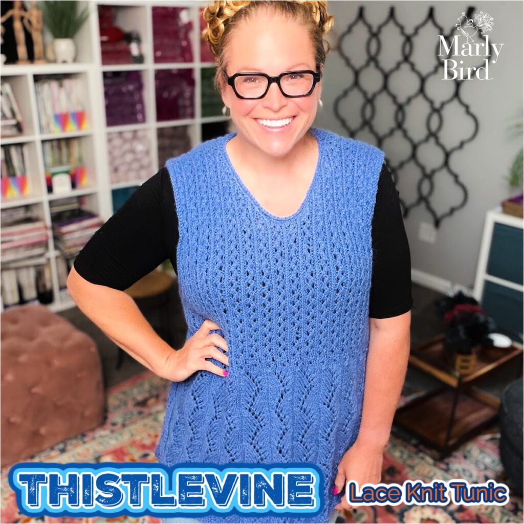 Woman models a blue lace knit tunic with intricate stitch detail, standing in a cozy room with shelves and decor behind her.