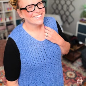 A woman models a hand-knit blue sleeveless vest, showing textured stitches and drape, in a cozy room with bookshelves.