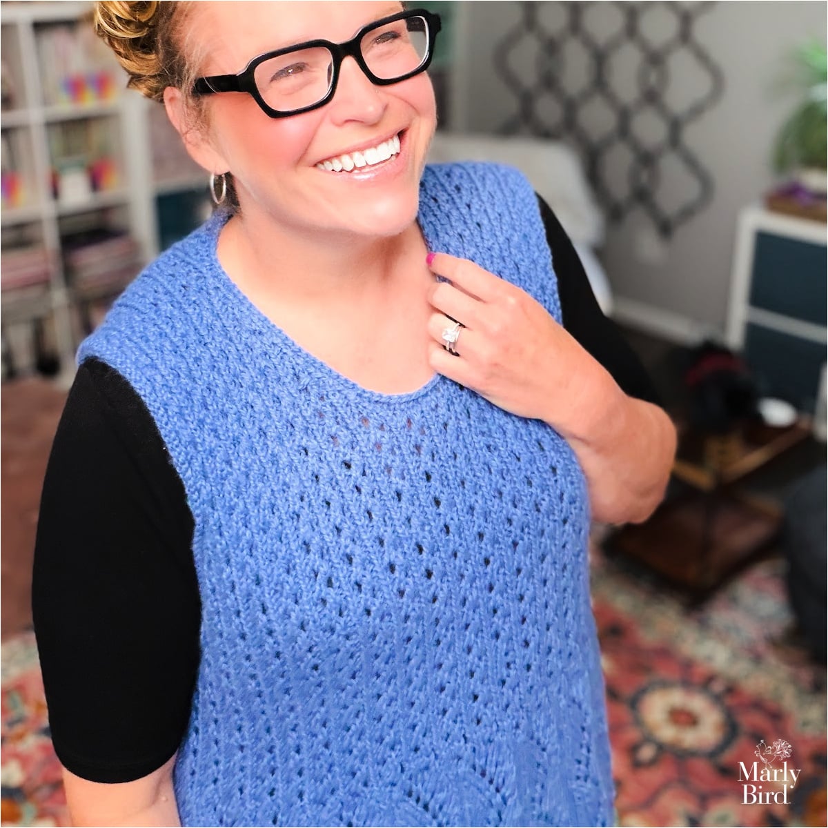 A woman models a hand-knit blue sleeveless vest, showing textured stitches and drape, in a cozy room with bookshelves.