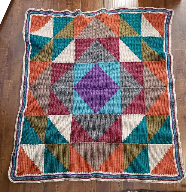 Crocheted blanket with triangles and squares in teal, orange, purple, beige, brown, olive; wavy striped edge on wood floor.